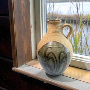 May include: A handmade ceramic jug in tan with a handle and spout. The lower half has a dark green and blue abstract pattern. It sits on a wooden windowsill, with a lake and reeds visible through the window.