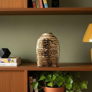 May include: A textured, brown and beige ceramic vase with a rounded shape and a narrow neck. The vase has horizontal lines and is displayed on a wooden shelf. The background includes books, a black geometric object, and a potted plant.