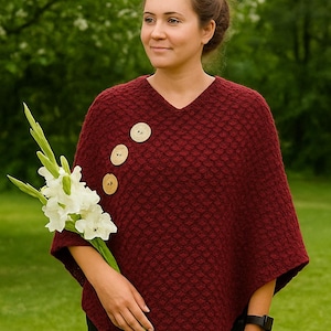 May include: A burgundy-colored, knitted poncho with a V-neck and three large wooden buttons. The poncho has a textured, diamond-like pattern. The person is holding white flowers with green stems. The background is a blurred green.