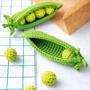 May include: Handmade crochet pea pods with bright green peas. Two pods are open, revealing the peas inside, while three individual peas are scattered nearby. The pods and peas are on a white surface with a green grid pattern.