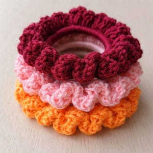 Könnte beinhalten: Ein Stapel von drei handgefertigten Häkel-Haargummis in Burgunderrot, Rosa und Orange. Die Haargummis haben ein gerüschtes, strukturiertes Design und sind übereinander gestapelt.