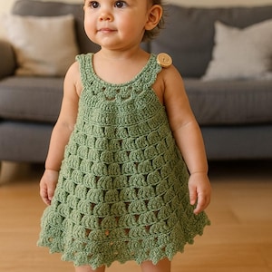 Può includere: Un vestito all'uncinetto verde con orlo smerlato e spalline, fissato con un bottone di legno. L'abito è indossato da una bambina. L'abito è di colore verde chiaro.