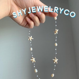 May include: A delicate necklace featuring a string of white pearls and five white starfish charms. The necklace is held by a hand against a light gray background. The text "SHY JEWELRY CO" is visible in the top right corner of the image.