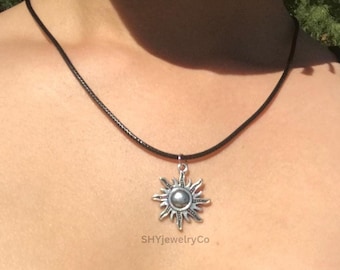 May include: A silver-colored sun pendant necklace with a black cord. The sun pendant has detailed rays and a central, possibly pearl-like, bead. The necklace is displayed against a person's skin, with a black top visible.