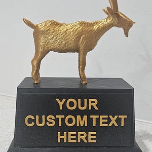 May include: A gold-colored goat trophy with a black base. The base features the text "YOUR CUSTOM TEXT HERE" in gold lettering. The goat figurine is detailed and stands on all four legs.