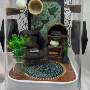 May include: Miniature diorama of a living room inside a clear, dome-shaped container. The scene includes a small armchair, bookshelf, rug, and artwork. Green plants and decorative elements add detail to the miniature interior.