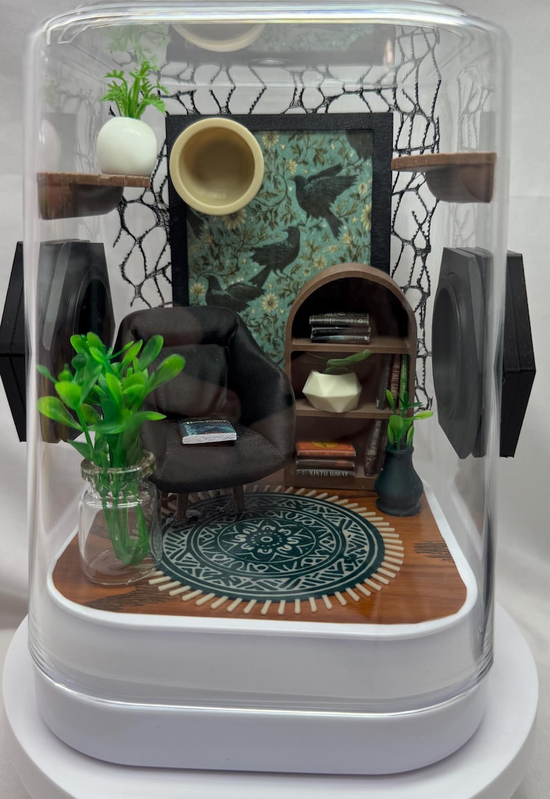 May include: Miniature diorama of a living room inside a clear, dome-shaped container. The scene includes a small armchair, bookshelf, rug, and artwork. Green plants and decorative elements add detail to the miniature interior.