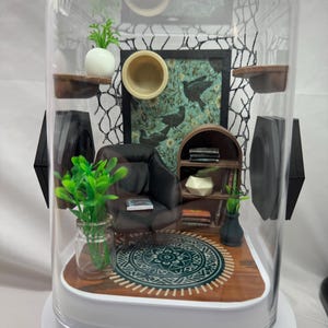 May include: A miniature diorama of a living room scene enclosed in a clear, rounded container. The scene includes a small armchair, a rug, a bookshelf, and decorative plants. A painting with birds hangs on the wall. The diorama is set on a white base.