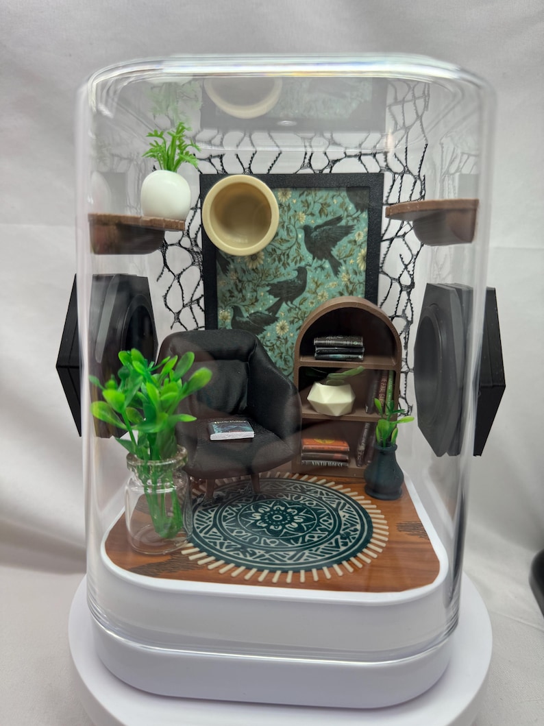 May include: A miniature diorama of a living room scene enclosed in a clear, rounded container. The scene includes a small armchair, a rug, a bookshelf, and decorative plants. A painting with birds hangs on the wall. The diorama is set on a white base.