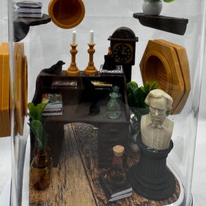 May include: Miniature diorama inside a clear, dome-shaped container. The scene includes a desk, bust, clock, candles, and other small items. The desk has books and a small plant. The bust is white, and the clock has a black face. The base has a wood-grain pattern.