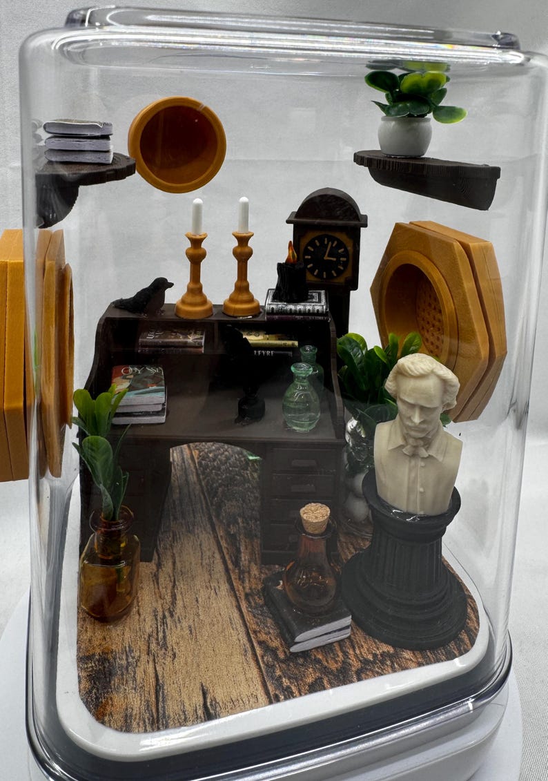May include: Miniature diorama inside a clear, dome-shaped container. The scene includes a desk, bust, clock, candles, and other small items. The desk has books and a small plant. The bust is white, and the clock has a black face. The base has a wood-grain pattern.