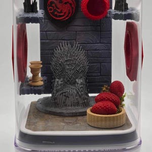 Può includere: Un diorama in miniatura del Trono di Spade di Game of Thrones, con un sigillo di drago rosso, una ciotola rossa e bacche rosse in un cestino di vimini. Il diorama è racchiuso in un cubo di plastica trasparente.