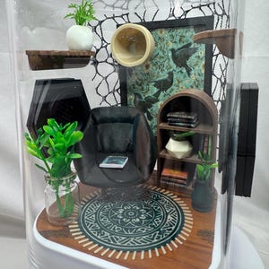 May include: Miniature interior scene inside a clear dome. The scene includes a black armchair, a patterned rug, a bookshelf, and decorative plants. A picture with birds hangs on the wall. The base is white and the interior is made of wood.