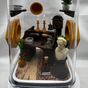 May include: Miniature diorama inside a clear display case. The scene includes a desk, bust, clock, candles, books, and plants. The diorama has a wooden floor and is enclosed in a clear, rectangular case with a white base.