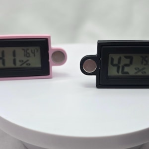 May include: Two digital hygrometers, one pink and one black, display temperature and humidity readings. Each device has a rectangular screen with rounded edges and magnetic attachments on either side.