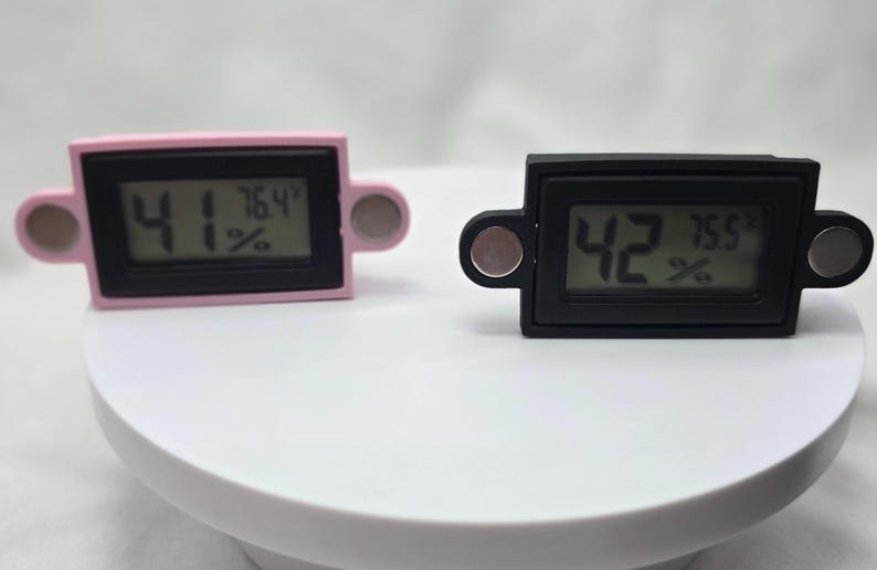 May include: Two digital hygrometers, one pink and one black, display temperature and humidity readings. Each device has a rectangular screen with rounded edges and magnetic attachments on either side.