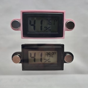 May include: Two digital thermometers, one pink and one black, displaying temperature and humidity readings. The pink thermometer shows 76.1&deg;F and 41% humidity. The black thermometer shows 75.2&deg;F and 41% humidity. Each has two magnetic attachments.