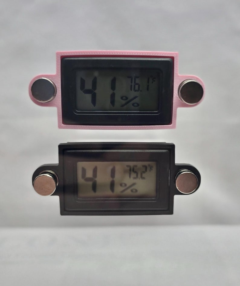May include: Two digital thermometers, one pink and one black, displaying temperature and humidity readings. The pink thermometer shows 76.1&deg;F and 41% humidity. The black thermometer shows 75.2&deg;F and 41% humidity. Each has two magnetic attachments.