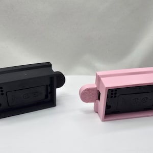 May include: Two rectangular, 3D-printed objects. One is black, the other is pink. Each has a rectangular body with a small, rounded handle on each side. The objects appear to be electronic device components.