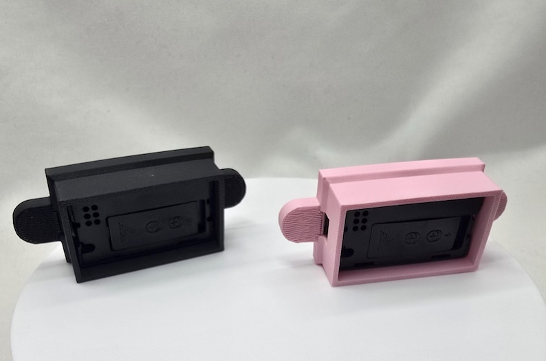 May include: Two rectangular, 3D-printed objects. One is black, the other is pink. Each has a rectangular body with a small, rounded handle on each side. The objects appear to be electronic device components.