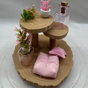 Puede incluir: Un diorama en miniatura con una estructura de madera de varios niveles con plantas artificiales y pequeños objetos decorativos. La escena incluye una pequeña almohada rosa, una concha y una botella de vidrio con un corcho.
