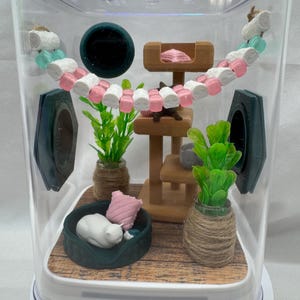 May include: A miniature cat-themed terrarium with a white cat figurine sleeping in a green bed, a wooden cat tree, and two potted plants. The terrarium is decorated with a pink and white garland.