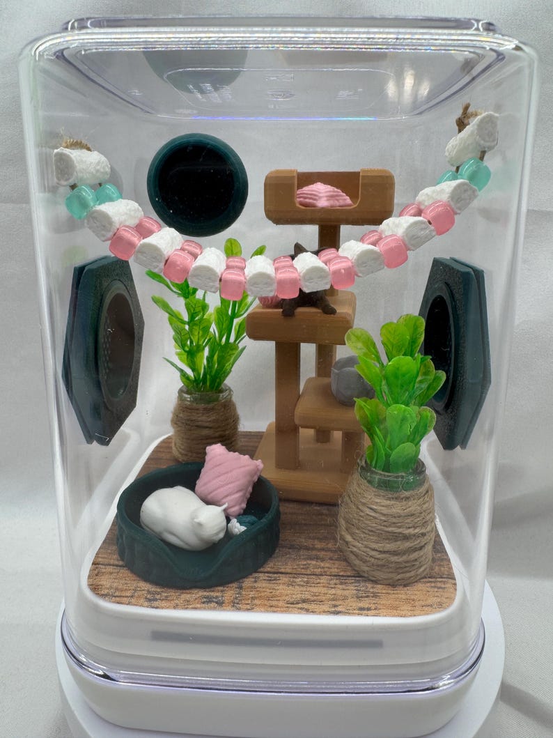 May include: A miniature cat-themed terrarium with a white cat figurine sleeping in a green bed, a wooden cat tree, and two potted plants. The terrarium is decorated with a pink and white garland.