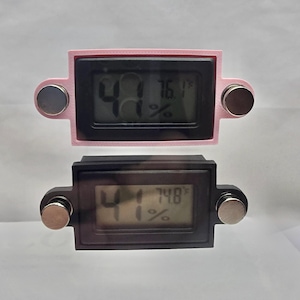 May include: Two digital hygrometers, one pink and one black, displaying humidity and temperature readings. Each device has a rectangular screen with a digital display and two silver magnets attached. The pink one reads 41% humidity and 76.1&deg;F.