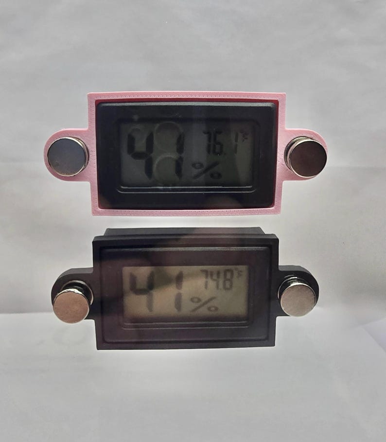 May include: Two digital hygrometers, one pink and one black, displaying humidity and temperature readings. Each device has a rectangular screen with a digital display and two silver magnets attached. The pink one reads 41% humidity and 76.1&deg;F.