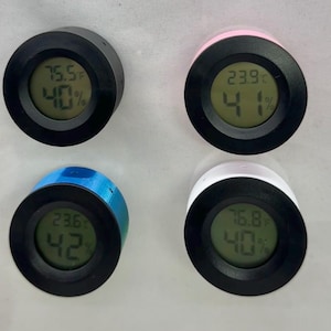 Puede incluir: Cuatro peque&ntilde;os higr&oacute;metros digitales redondos con caras negras y bordes de colores. Cada uno muestra la temperatura en grados Fahrenheit y el porcentaje de humedad. Los bordes son negros, rosas, azules y blancos. Las pantallas muestran diferentes temperaturas y niveles de humedad.