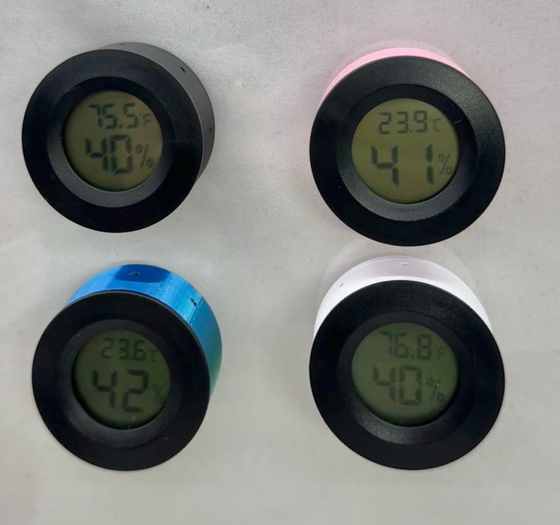Puede incluir: Cuatro peque&ntilde;os higr&oacute;metros digitales redondos con caras negras y bordes de colores. Cada uno muestra la temperatura en grados Fahrenheit y el porcentaje de humedad. Los bordes son negros, rosas, azules y blancos. Las pantallas muestran diferentes temperaturas y niveles de humedad.