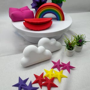 Può includere: Una collezione di oggetti decorativi colorati stampati in 3D. Include un arcobaleno, stelle viola, rosse e gialle, nuvole bianche, un pouf rosa e una stella geometrica viola. Inoltre, piccole piante in vaso.