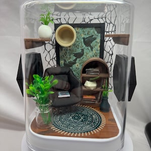 May include: Miniature diorama of a living room scene inside a clear display case. The scene includes a small black armchair, a patterned rug, a bookshelf, and decorative plants. The background features a framed picture with bird illustrations.