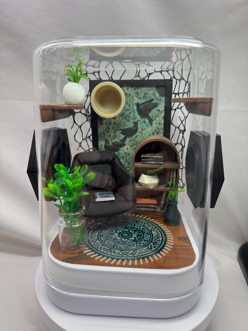 May include: Miniature diorama of a living room scene inside a clear display case. The scene includes a small black armchair, a patterned rug, a bookshelf, and decorative plants. The background features a framed picture with bird illustrations.
