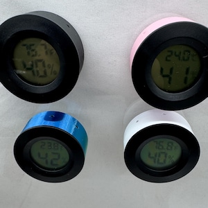 Puede incluir: Cuatro peque&ntilde;os higr&oacute;metros digitales redondos en negro, rosa, azul y blanco. Cada dispositivo tiene un bisel negro y muestra la temperatura en grados Celsius y el porcentaje de humedad en una pantalla digital.