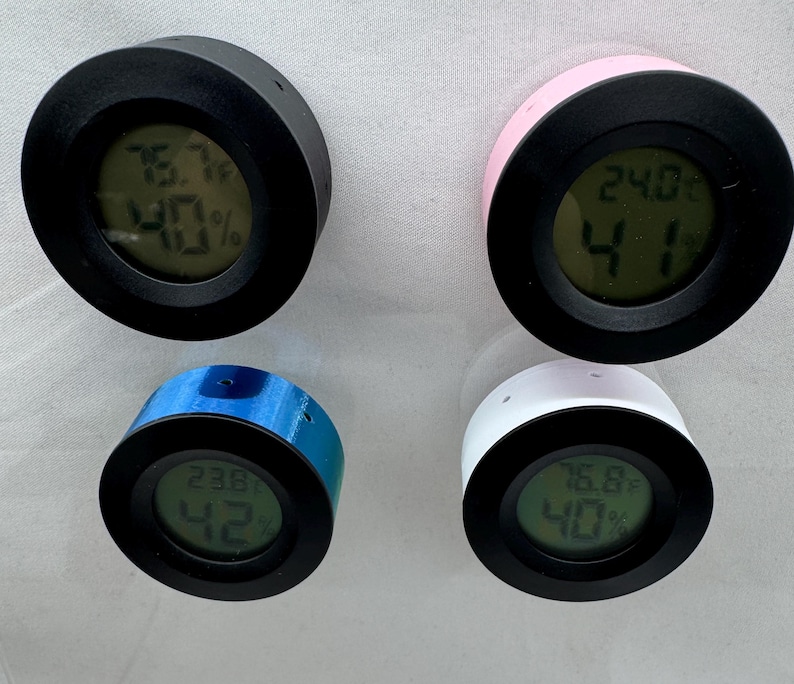 Puede incluir: Cuatro peque&ntilde;os higr&oacute;metros digitales redondos en negro, rosa, azul y blanco. Cada dispositivo tiene un bisel negro y muestra la temperatura en grados Celsius y el porcentaje de humedad en una pantalla digital.