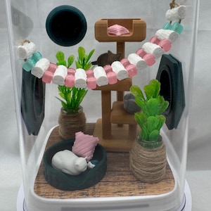May include: A miniature cat-themed diorama inside a clear plastic container. The diorama features a white cat sleeping in a bed, a cat tree with a pink cushion, a green plant, and a pink and white beaded cat toy.
