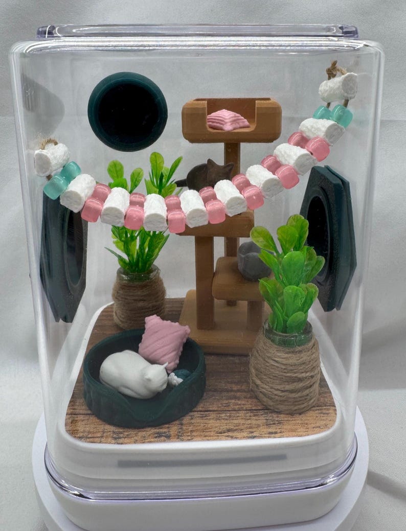 May include: A miniature cat-themed diorama inside a clear plastic container. The diorama features a white cat sleeping in a bed, a cat tree with a pink cushion, a green plant, and a pink and white beaded cat toy.