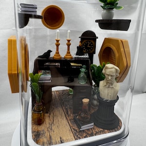 May include: A miniature diorama inside a clear, rectangular display case. The scene includes a desk, bust, clock, candles, and plants. The desk has books and a small bottle. The bust is white, and the clock is black.