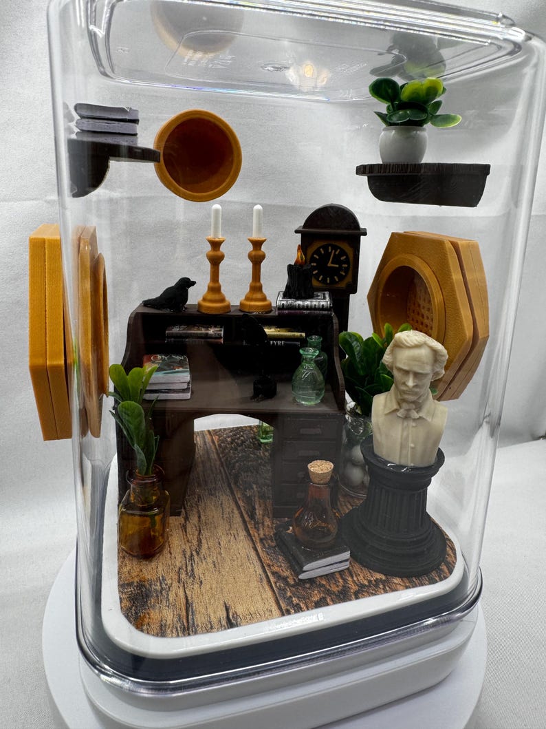 May include: A miniature diorama inside a clear, rectangular display case. The scene includes a desk, bust, clock, candles, and plants. The desk has books and a small bottle. The bust is white, and the clock is black.