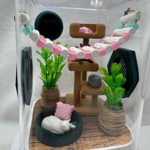 May include: A miniature cat house with a white cat figurine sleeping in a green bed, a wooden cat tree, and two potted plants. The house is inside a clear plastic container with a wooden floor.