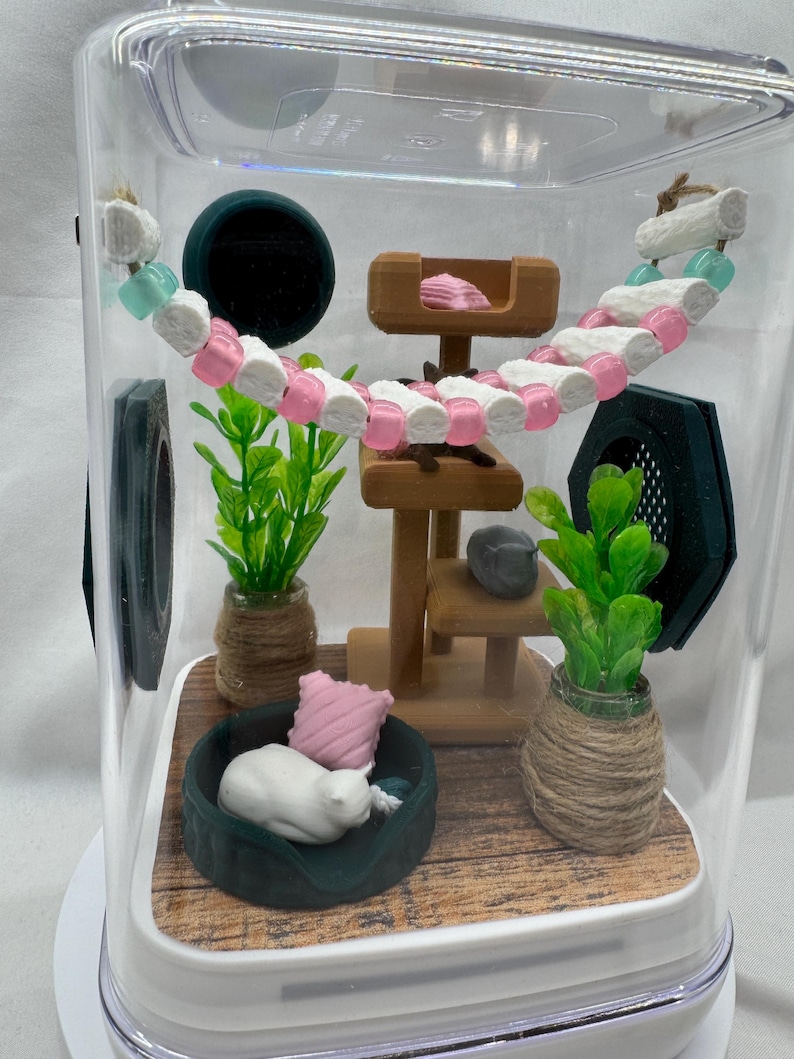 May include: A miniature cat house with a white cat figurine sleeping in a green bed, a wooden cat tree, and two potted plants. The house is inside a clear plastic container with a wooden floor.