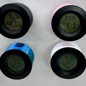 Puede incluir: Cuatro peque&ntilde;os higr&oacute;metros digitales redondos en negro, rosa, azul y blanco. Cada uno muestra la temperatura en grados Celsius y el porcentaje de humedad en una pantalla digital verde. El higr&oacute;metro negro muestra 24,3&deg;C y 40% de humedad.