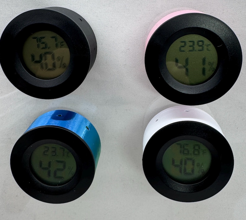 Puede incluir: Cuatro peque&ntilde;os higr&oacute;metros digitales redondos en negro, rosa, azul y blanco. Cada uno muestra la temperatura en grados Celsius y el porcentaje de humedad en una pantalla digital verde. El higr&oacute;metro negro muestra 24,3&deg;C y 40% de humedad.