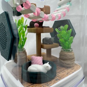 May include: A miniature cat tree with a pink and white cat bed, a white cat figurine, and green plants in a clear plastic container.