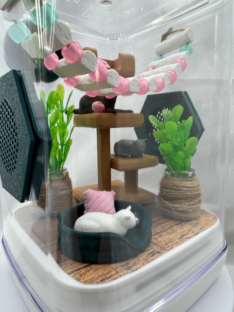 May include: A miniature cat tree with a pink and white cat bed, a white cat figurine, and green plants in a clear plastic container.