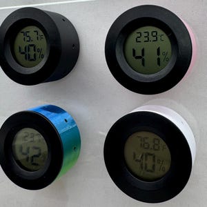 Puede incluir: Cuatro peque&ntilde;os term&oacute;metros digitales redondos con bordes negros y cuerpos de varios colores: negro, rosa, degradado azul-verde y blanco. Cada uno muestra la temperatura en grados Celsius y el porcentaje de humedad en una pantalla verde. Montados sobre una superficie gris claro.