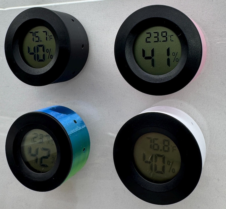 Puede incluir: Cuatro peque&ntilde;os term&oacute;metros digitales redondos con bordes negros y cuerpos de varios colores: negro, rosa, degradado azul-verde y blanco. Cada uno muestra la temperatura en grados Celsius y el porcentaje de humedad en una pantalla verde. Montados sobre una superficie gris claro.