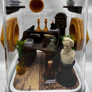 May include: Miniature diorama inside a clear dome. The scene includes a desk, bust, clock, candles, and books. The desk is dark brown, and the bust is white. The dome sits on a white base, and the background is a light gray.