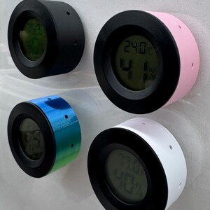 Puede incluir: Cuatro peque&ntilde;os higr&oacute;metros digitales redondos en negro, rosa, azul-verde y blanco. Cada uno tiene un bisel negro y una pantalla digital que muestra la temperatura y la humedad. Los higr&oacute;metros est&aacute;n montados sobre una superficie blanca.